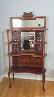 Front view of the full vintage Victorian mahogany curio stand showing top mirror, carved cresting, shelves and carved side panels.