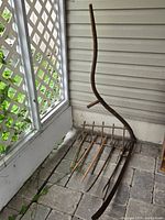 Photo of vintage hay sickle positioned upright against a wall showing the full length including wooden handle and rusted metal prongs.