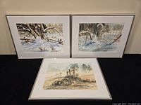 Three framed watercolour paintings, two in vertical orientation showing snow and trees, one horizontal showing rural field.