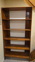 Front view of tall wooden bookcase with five shelves, shown empty, placed against a wall near a staircase. The wood finish is medium brown with visible grain.
