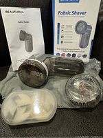 Fabric shaver device with user manual, original box, additional blade heads and protective covers visible on gray fabric.