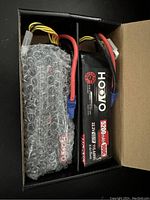 Two Hoovo branded 5200mAh 22.2V 100C Li-po batteries side by side in a cardboard box, one partly wrapped in bubble wrap with multiple wires and blue connectors