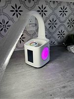 White flexible-neck LED desk lamp with cube-shaped base, side multi-color LED light showing pink, digital color wheel, pen storage, USB port visible in base corner.