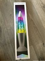 Box front showing West & Arrow Glitter Lamp with rainbow multi-color glitter and metallic silver base on wooden floor.