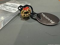 Close-up photo of the Pandora pig charm in gold with red enamel bow, showing the attached black string and round Pandora tag, lying next to original sealed plastic packaging.