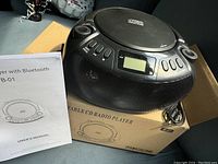 Photo of the portable CD radio player on its box with user manual visible, showing control buttons, display, and speaker grille.