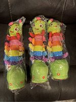 Three new plush rainbow-colored singing dancing caterpillar toys sealed in individual plastic bags, showing their full body and colors.