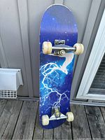 Front view of skateboard showing blue deck with white lightning and dragon graphic, white wheels, metal trucks.