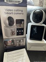 Front view of video calling smart camera on stand with display screen, original box and instruction sheet showing product features.