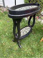 Black oval wood table with glass top and lower shelf placed on grass. Visible signs of wood finish wear and chipping.