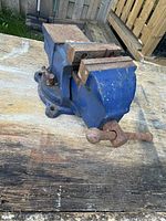 Blue metal work bench vice on wooden surface showing rust and wear on handle and jaws.