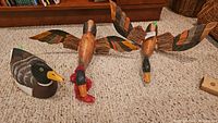 Three wooden duck figurines on a carpet; two appear airborne with patterned wings, one a standing mallard decoy style duck.