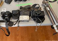 Overview of the lot with 3 digital cameras, 1 film camera, and tripod on a table