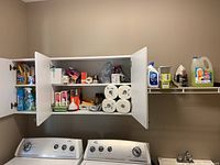View of opened wall cabinet above washer and dryer showing assorted cleaning supplies including sprays, liquids, paper towels, light bulbs, and other household items.