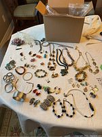 Photo showing assortment of costume jewelry including multiple necklaces, earrings, bracelets, and partial pieces spread on table with box in background.