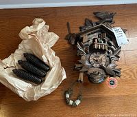 Cuckoo clock with three pinecone weights wrapped in paper laid beside it on the floor with a tag visible.