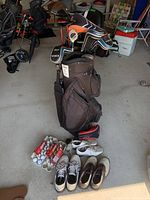 Full view of golf bag filled with clubs, multiple golf balls, and three pairs of golf shoes on the floor