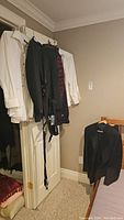 Two white dress shirts, black tuxedo jackets, matching tuxedo pants, red and black patterned cummerbunds and bow ties hanging from door hooks and on a chair.