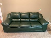 Front view of a three-seater green leather sofa with wide armrests and cushioned backrests, positioned on carpeted floor against a beige wall.