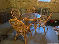 Round rattan table with glass top and four matching rattan chairs around it, placed in a basement with concrete floor and unfinished walls.