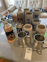 Image showing a collection of 10 beer steins arranged on a table, including ceramic and metal varieties with various designs and sizes.