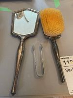 Full view of sterling silver hand mirror, hair brush with natural bristles, and silver tongs on tabletop
