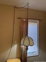 View of hanging swag lamp with wicker scalloped dome shade suspended by twisted chain in a room with window and curtain.