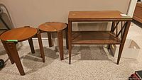 Three wood side tables arranged on carpet: two round matching tables and one rectangular two-tier table.