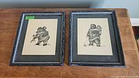 Two framed Inuit prints side by side on a wooden table, showing figures carrying children and holding fish, signed Hilda Laurie.