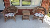 Set of two brown painted wicker chairs with floral cushions and one matching ottoman on outdoor porch.