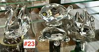 Four clear crystal animal figurines displayed on glass shelf include two swans, one turtle, and one fish, with faceted cuts and reflective properties.