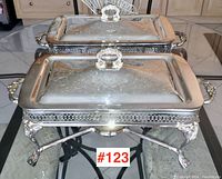 Two silver plated rectangular serving caddies with lids resting on glass table, showing top views and ornate handles.