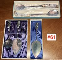 Six serving pieces including two porcelain-handled salad servers in a box and three silver-plated servers with rose-shaped handles in silk-lined boxes, all arranged on a wooden surface.