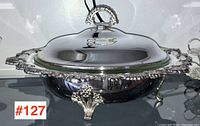 Front angled view of silver plated serving dish with domed lid and ornate handle, showing reflective surface and decorative feet.
