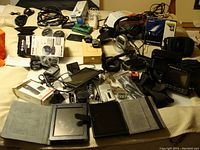 Full view of assorted electronics and gadgets spread on a surface including cameras, tablets, GPS units, headphones, and cables.