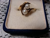 Front view showing two white pearls mounted in a sculptural 10K gold setting, in a cream velvet ring box with blue trim.
