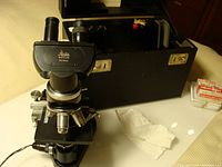 Full view of microscope on stage with original black case open behind it and slide pack visible.