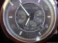 Close-up of the watch dial showing the Michael Kors brand, chronograph subdials, and date window at 4 o'clock position. The dial has a black background with silver-tone markers and hands. The crystal has a visible scratch.