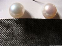 Close-up photo of two round pearl stud earrings with a white backing card, showing smooth surfaces and natural pearl luster. The posts appear metallic and are marked 10K gold.