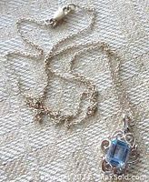 Full view of necklace chain and pendant showing delicate silver cable oval link chain and lobster clasp alongside small blue glass stone pendant in openwork frame.