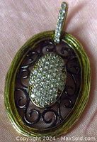 Close-up front view of oval pendant showing diamond paved center, dark scrollwork, and textured gilded silver outer frame with diamond accented bail.