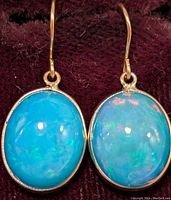 Close-up of the pair of gold earrings featuring oval enhanced blue opal stones against a dark fabric background.
