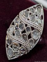 Close-up of marquise-shaped stainless steel ring with multiple pear-shaped diamonds and wavy bands of smaller diamonds over open lattice metalwork, showing detailed design on ring surface.