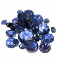 Close-up top-view photo of multiple loose faceted sapphire gemstones in various sizes and cuts, displaying deep blue to violet hues with reflective facets.