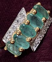 Close-up view of the ring showing five oval green emerald stones and white topaz stones on triangular silver panels.