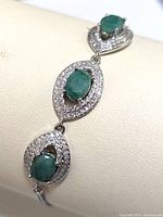 Close-up view of three oval green emerald stones set in silver with two rows of small white cubic zirconia stones surrounding each stone.