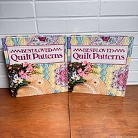Two binders titled 'Best Loved Quilt Patterns' standing side by side on wood surface against white brick wall.