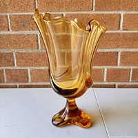 Front angled view of vintage MCM Viking amber pedestal glass vase with waved rim and three-toe base, showing the warm amber color and clear glass highlights.