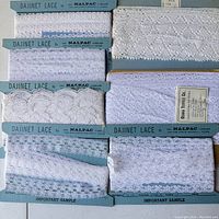 Eight vintage white lace trims each attached to a blue card labeled 'DAJINET LACE by NALPAC' showing different lace patterns and textures.