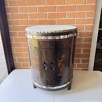 Closed view of the oriental themed wooden drum table cabinet showing carved figures and metal studded rims.